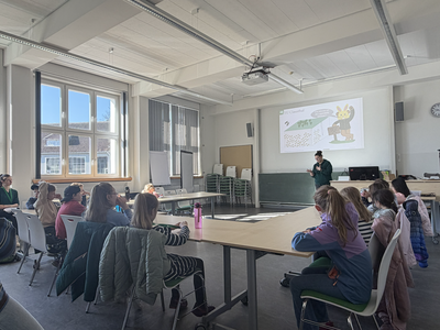 rontale Ansicht auf eine Dozentin, die vor einer Sitzgruppe von aufmerksam zuhörenden Kindern in einem Seminarraum steht und den Lehrstoff mit Gestik unterstreicht. Die Präsentationsfolie im Hintergrund visualisiert die Leitfrage „Wie können wir die Lagerstätte abbauen?“ anhand eines geologischen Blockbildes und eines Osterhasen in Bergmannskluft. Die Aufnahme zeigt die Erarbeitung der theoretischen Konzepte zur Bergwerksplanung.