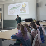 rontale Ansicht auf eine Dozentin, die vor einer Sitzgruppe von aufmerksam zuhörenden Kindern in einem Seminarraum steht und den Lehrstoff mit Gestik unterstreicht. Die Präsentationsfolie im Hintergrund visualisiert die Leitfrage „Wie können wir die Lagerstätte abbauen?“ anhand eines geologischen Blockbildes und eines Osterhasen in Bergmannskluft. Die Aufnahme zeigt die Erarbeitung der theoretischen Konzepte zur Bergwerksplanung.