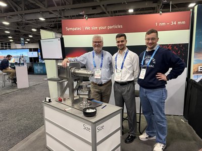 Drei Männer stehen zusammen an einem Messestand der Firma Sympatec. Auf dem Tresen vor ihnen ist ein technisches Messgerät platziert. Die Person auf der rechten Seite trägt einen dunkelblauen Pullover mit der Aufschrift „Technische Universität Clausthal“.