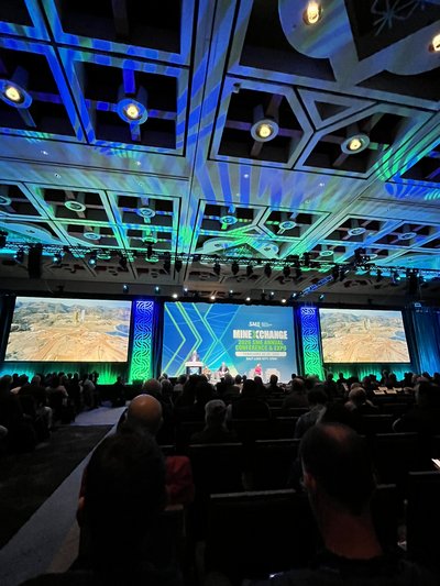 Blick aus dem Publikum auf die Bühne eines großen Vortragssaals mit blauer und grüner Deckenbeleuchtung. Auf der Bühne findet eine Podiumsdiskussion statt. Die mittlere Leinwand zeigt den Schriftzug „SME MINEXCHANGE 2026“, auf den seitlichen Leinwänden ist ein Bergbaugebiet zu sehen.