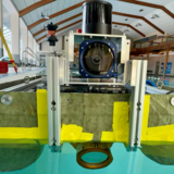 Kastenförmige, schwimmende Plattform TB1 im hellen Hallenbad, frontal aufgenommen: In der Mitte ein großes, rundes Kamera‑/Sensorsystem in einem Metallrahmen, darunter der Bug knapp über der Wasseroberfläche. Links hinten am Beckenrand steht ein gelbes Tachymeter auf rotem Stativ als Referenzmesssystem.