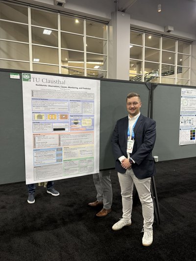 Ein Konferenzteilnehmer im dunklen Sakko steht vor seinem großen wissenschaftlichen Poster. Das Poster trägt das Logo der TU Clausthal und den Titel „Rockbursts: Observations, Causes, Monitoring, and Prediction“.