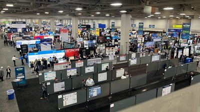 Ein Blick von oben in eine weitläufige, belebte Messehalle. Im Vordergrund befindet sich ein Bereich mit zahlreichen wissenschaftlichen Postern auf grauen Stellwänden, im Hintergrund reihen sich die beleuchteten Stände verschiedener Industrieunternehmen aneinander.