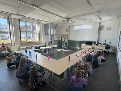 Ein heller Seminarraum mit u-förmig angeordneten Tischen, neben denen mehrere Kinder und eine Dozentin tief in der Hocke auf dem Boden verweilen. Im Hintergrund projiziert ein Beamer das Logo der TU Clausthal, die Worte „Glück auf, wenn du…“ sowie das Bergbau-Symbol Schlägel und Eisen an die Wand. Die Szene dokumentiert ein interaktives Kennenlernspiel zur Einführung in die bergbaulichen Grundlagen.