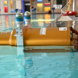 In einem Schwimmbad- bunter Hintergrund, blaues Wasser. IM Vordergrund ein oranger Zylinder