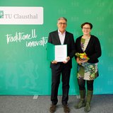Ein Mann im dunklen Anzug und eine Frau in buntem Kleid mit schwarzer Jacke stehen frontal nebeneinander vor einer grünen Wand mit TU-Clausthal-Logo und dem Schriftzug „traditionell innovativ“. Der Mann hält eine geöffnete Verabschiedungsurkunde in den Händen, die Frau einen kleinen Blumenstrauß. Die Szene zeigt die offizielle Verabschiedung von Prof. Hossein Tudeshki durch Universitätspräsidentin Sylvia Schattauer nach seinem langjährigen Wirken im Tagebau und internationalen Bergbau.