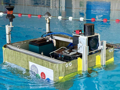 TB1 während der Fahrt im Schwimmbecken: Die rechteckige Plattform mit Sensorik und Kabeln erzeugt sichtbare Wellen an der Wasseroberfläche. Am Heck ragen zwei schlanke Masten mit Prismas und kleiner Sensorik nach oben, seitlich ist erneut das Projektlogo zu erkennen; im Hintergrund verlaufen weiß‑rote Schwimmleinen entlang des Beckens.