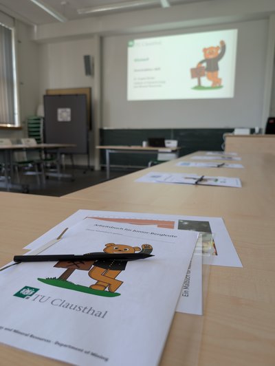 Im Vordergrund ein gehefteter Stapel Papier mit dem Titel Arbeitsbuch für Junior-Bergleute, darunter ein winkender Bär im Bergkittel und darunter eine TU Clausthal Logo. Abgeschnitten darunter steht Institute of Geotechnology and Mineral Resources - Department of Mining. Im Hintergrund unscharf eine Wand mit einer Projektion des winkenden Bären auf einer Folie im TU Design