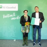 in Mann und eine Frau stehen frontal nebeneinander vor einer grünen Wand mit dem Logo der TU Clausthal und dem Schriftzug „traditionell innovativ“. Die Frau in gemusterter Kleidung hält einen kleinen Blumenstrauß in der Hand, der Mann in Sakko und Jeans präsentiert eine geöffnete Verabschiedungsurkunde. Die Szene zeigt die offizielle Verabschiedung des Geotechnik-Professors Norbert Meyer durch Universitätspräsidentin Sylvia Schattauer.