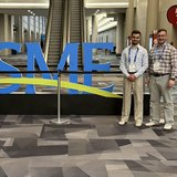 Zwei Konferenzteilnehmer stehen lächelnd neben einem großen, blauen 3D-Schriftzug mit den Buchstaben „SME“ im Foyer eines Kongresszentrums. Im Hintergrund sind Rolltreppen zu sehen.