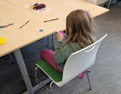 Ein Kind, das etwas aus pinkem Karton ausschneidet, auf einem Stuhl sitzend von hinten fotographiert